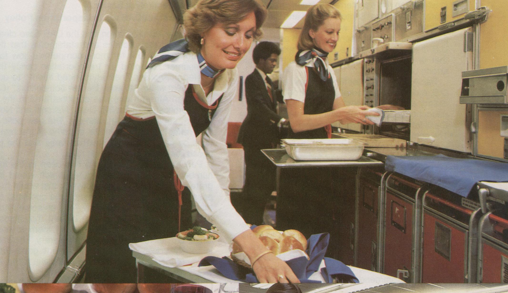 1977,  Flight Attendants preparing to serve an elaborate First Class meal in the galley of a Pan Am Boeing 747SP.  Sue Smith is preparing the serving cart.  Judy Skartvedt is standing by the open oven and Antonio Gooding is working at the back of the galley.  